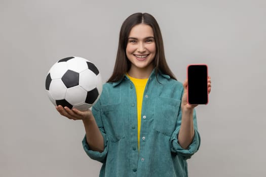 Smiling positive woman with dark hair holding and showing blank display smartphone and football ball, betting on soccer, wearing casual style jacket. Indoor studio shot isolated on gray background.