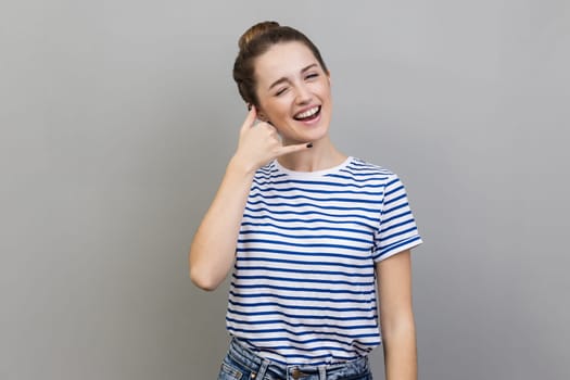 Call me back. Portrait of woman wearing striped T-shirt standing with happy playful expression and making telephone gesture with hand flirting. Indoor studio shot isolated on gray background.