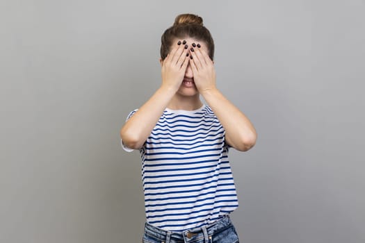 Don't want to look at this. Portrait of confused woman wearing striped T-shirt covering eyes with hand, feeling shamed and scared to watch. Indoor studio shot isolated on gray background.