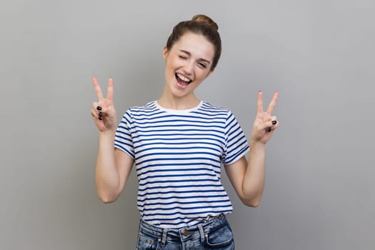 Portrait of happy successful woman wearing striped T-shirt showing v sign victory gesture with fingers, looking at camera with smile. Indoor studio shot isolated on gray background.