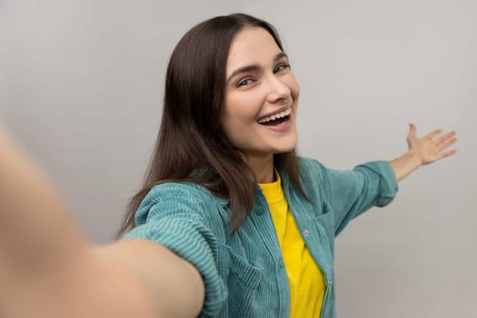 Portrait of smiling happy woman making point of view photo, showing welcome gesture, positive emotions, wearing casual style jacket. Indoor studio shot isolated on gray background.