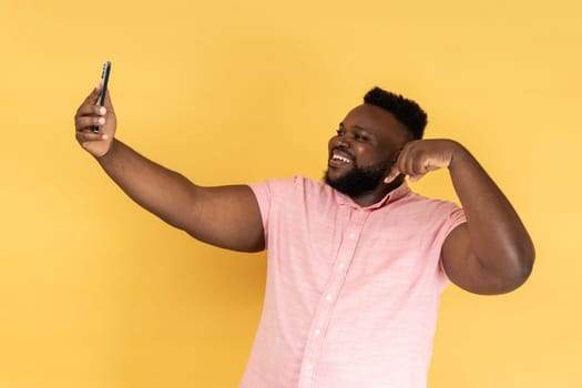 Portrait of handsome positive man wearing pink shirt taking selfie or having livestream, pointing finger down, subscribe his vlog. Indoor studio shot isolated on yellow background.