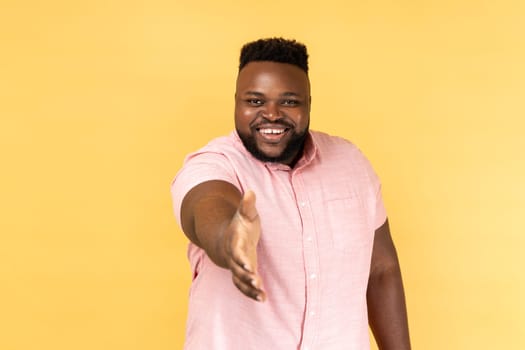 Hello, nice to meet you. Portrait of man in pink shirt giving hand to handshake and smiling friendly, consultant or manager offering job, partnership. Indoor studio shot isolated on yellow background.