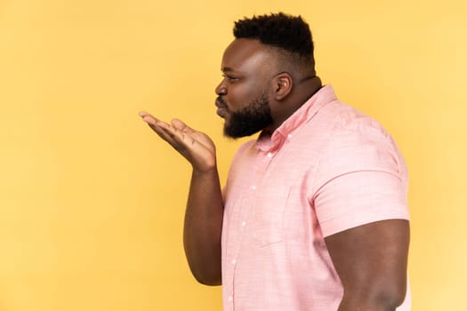 Side view portrait of falling in love young adult man wearing pink shirt sending air kissing over palms, expressing romantic feelings, flirting. Indoor studio shot isolated on yellow background.