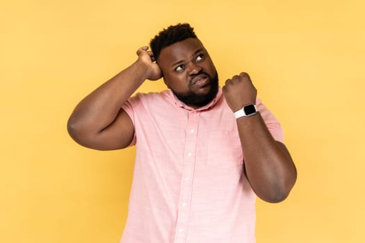 Portrait of puzzled confused man wearing pink shirt showing his smart watch and scratching his head, looking away with with thoughtful expression. Indoor studio shot isolated on yellow background.