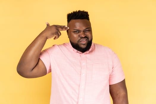 Kill me pleas. Portrait of desperate man wearing pink shirt pointing finger gun to head, shooting herself, making suicide gesture to stop depression. Indoor studio shot isolated on yellow background.