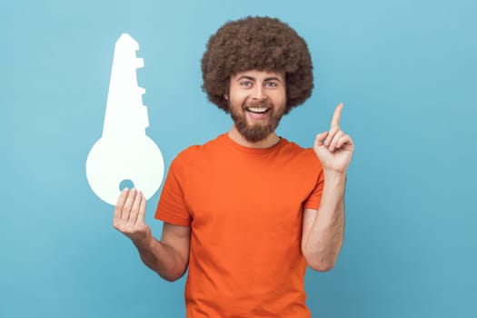 Portrait of smiling man with Afro hairstyle wearing orange T-shirt holding big paper key and pointing finger up, having idea for renting new flat. Indoor studio shot isolated on blue background.