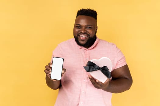 Portrait of joyful cheerful bearded man wearing pink shirt holding heart shaped present box and showing mobile phone with empty display. Indoor studio shot isolated on yellow background.