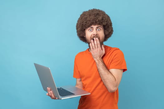 Portrait of man with Afro hairstyle wearing orange T-shirt working on notebook, sees something astonishing, covering mouth with palm. Indoor studio shot isolated on blue background.