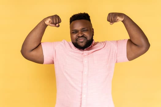 Portrait of strong and independent self confident man wearing pink shirt showing biceps raising strong arms, proud of muscular build, leadership. Indoor studio shot isolated on yellow background.