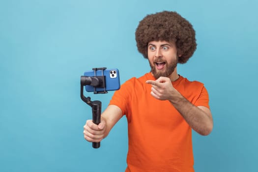 Portrait of astonished man with Afro hairstyle wearing orange T-shirt using stedicam and phone for livestream, being amazed, pointing at phone camera. Indoor studio shot isolated on blue background.