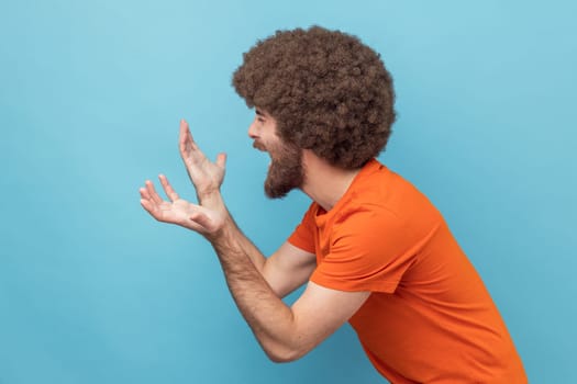 Side view of man with Afro hairstyle wearing orange T-shirt spreading hands, seriously looking at camera, family quarrel, misunderstanding. Indoor studio shot isolated on blue background.