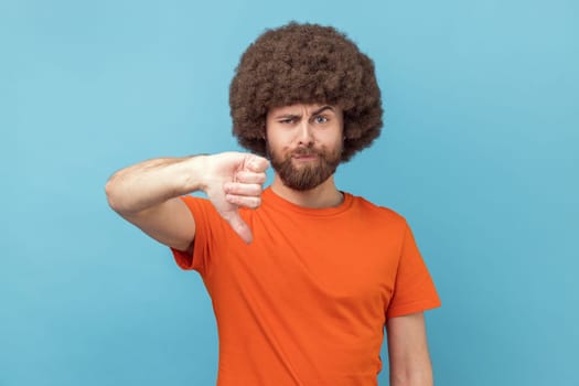 Portrait of displeased man with Afro hairstyle wearing orange T-shirt showing thumbs down dislike gesture, symbol of disagree, giving feedback. Indoor studio shot isolated on blue background.