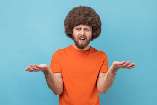 Portrait of stressed annoyed man with Afro hairstyle wearing orange T-shirt standing raised arms and asking, expressing negative emotions. Indoor studio shot isolated on blue background.