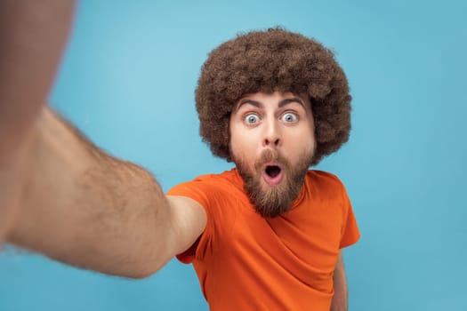 Portrait of surprised shocked man with Afro hairstyle wearing orange T-shirt taking selfie or broadcasting livestream, looking with wow expression, POV. Indoor studio shot isolated on blue background.