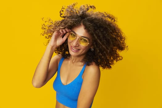 Woman with curly afro hair in a blue T-shirt on a yellow background dancing flying hair with sunglasses yellow, hand signs, look into the camera, smile with teeth and happiness, copy space. High quality photo