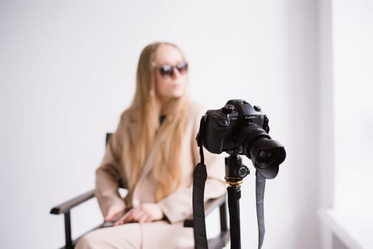 A woman videographer in sunglasses, a blonde with a camera on a tripod in the studio. Wearing a formal nude pantsuit on a white isolated background, person defocus