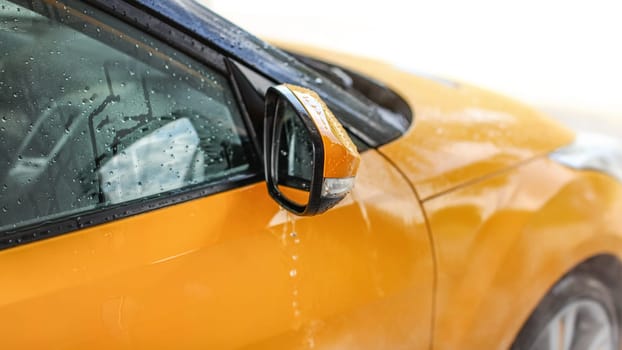 Water dripping from wet side mirror of yellow car washed in carwash.