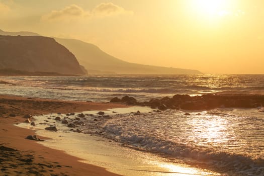 Sunset on empty wild beach. Small waves over rock, hills in hazy background.