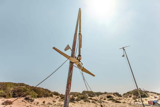 Two small wind turbines, one of them broken, standing on desert, strong back light sun in background.