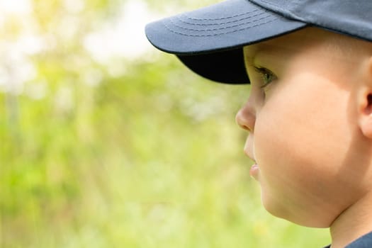 Face in profile of a little handsome boy in a blue cap on a green background in the sun. Close-up. copy space. Banner.