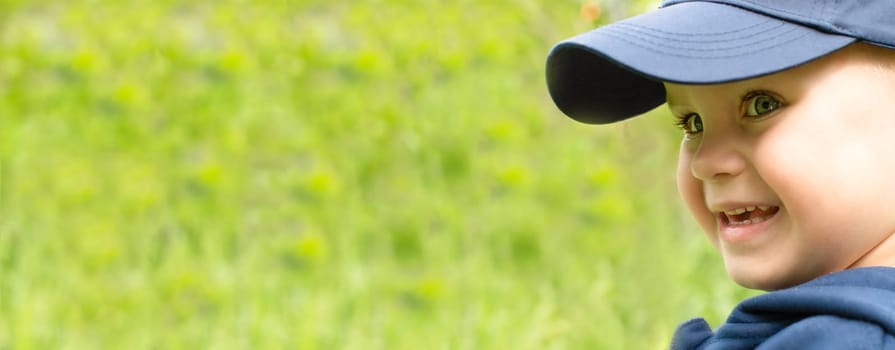 Face in profile of a little cheerful and handsome boy in a blue cap, smiling happily on a green background. close-up. Banner. copy space.