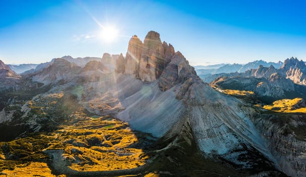 Landscape of rocky Three Peaks of Lavaredo at sunrise. Curved roads with cars on highland slopes around mountain under blue sky aerial view
