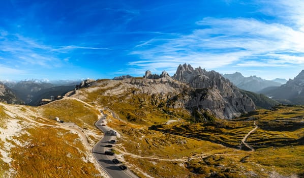 Road winds at foot of rocky mountain in Tre Cime di Lavaredo. Scenic mountain ranges under blue sky with white clouds at sunset aerial view
