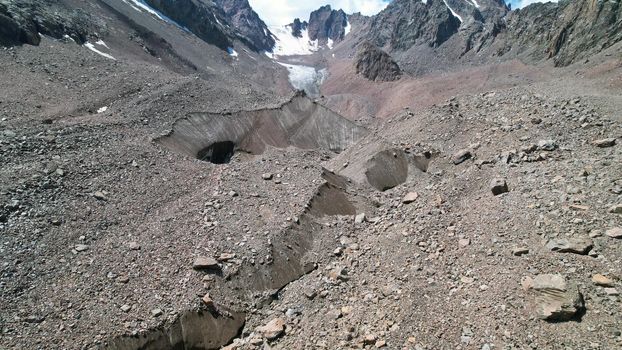 A moraine lake among a glacier covered with rocks. The glacier is melting. Shadows from clouds. View of the lake from above from the throne. A lake among high snowy mountains. Kazakhstan, Almaty