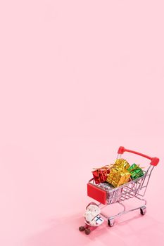 Christmas shopping concept, mini red shop cart trolley with Santa Claus toy and gift box isolated on pale pink background, blank copy space, close up
