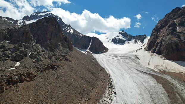 High rocky mountains are sometimes covered with ice and snow. A huge glacier passes between the peaks. The ice is gradually melting. The stones are lying on glacier. Steep cliffs. The view from drone