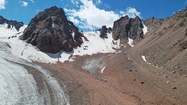 High rocky mountains are sometimes covered with ice and snow. A huge glacier passes between the peaks. The ice is gradually melting. The stones are lying on glacier. Steep cliffs. The view from drone