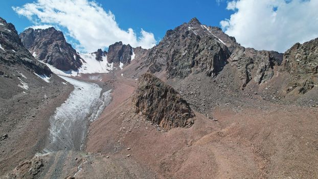 High rocky mountains are sometimes covered with ice and snow. A huge glacier passes between the peaks. The ice is gradually melting. The stones are lying on glacier. Steep cliffs. The view from drone