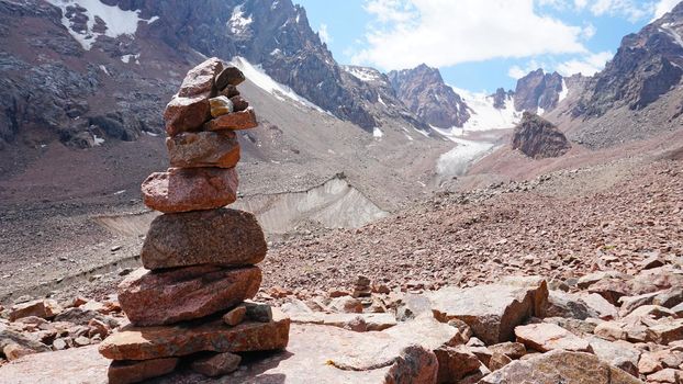 A pyramid of stones as a pointer in the mountains. Big rocks, high peaks and a snowy glacier. A moraine lake can be seen in the distance. The ice is covered with huge rocks. A cairn.