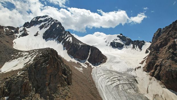 High rocky mountains are sometimes covered with ice and snow. A huge glacier passes between the peaks. The ice is gradually melting. The stones are lying on glacier. Steep cliffs. The view from drone