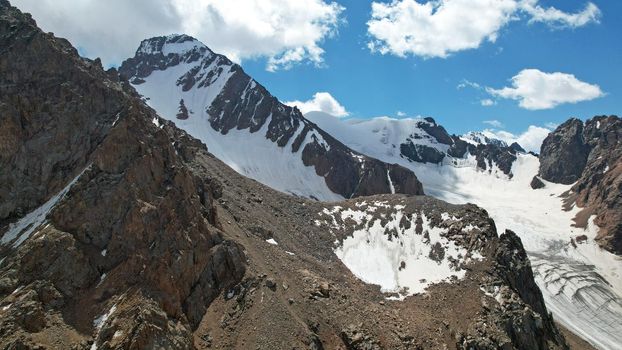 High rocky mountains are sometimes covered with ice and snow. A huge glacier passes between the peaks. The ice is gradually melting. The stones are lying on glacier. Steep cliffs. The view from drone