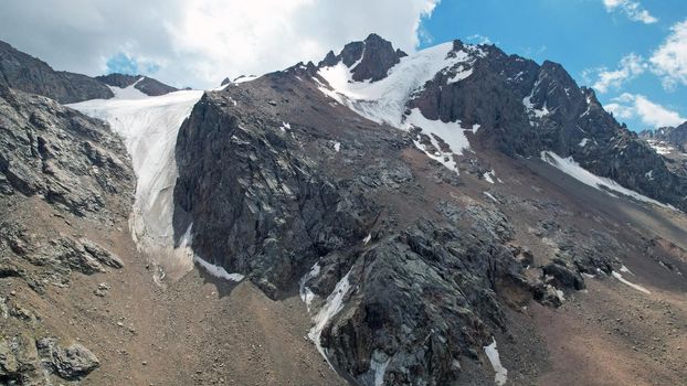 High rocky mountains are sometimes covered with ice and snow. A huge glacier passes between the peaks. The ice is gradually melting. The stones are lying on glacier. Steep cliffs. The view from drone