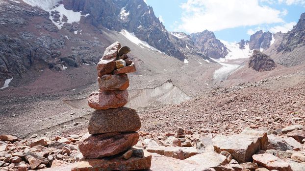 A pyramid of stones as a pointer in the mountains. Big rocks, high peaks and a snowy glacier. A moraine lake can be seen in the distance. The ice is covered with huge rocks. A cairn.