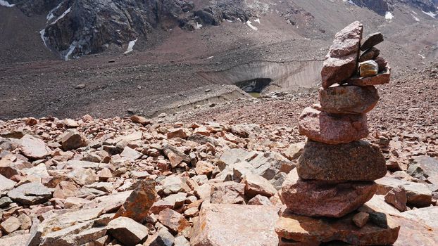 A pyramid of stones as a pointer in the mountains. Big rocks, high peaks and a snowy glacier. A moraine lake can be seen in the distance. The ice is covered with huge rocks. A cairn.