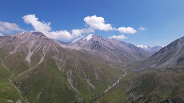 Clouds flow around the tops of high mountains. Rocky gorges and cliffs. The sky is blue, the sun is shining brightly, green grass is growing in places. Mountain terrain from the height of the drone