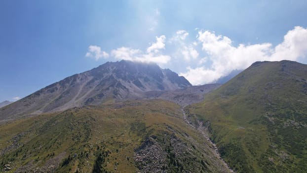 Clouds flow around the tops of high mountains. Rocky gorges and cliffs. The sky is blue, the sun is shining brightly, green grass is growing in places. Mountain terrain from the height of the drone