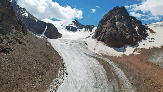 High rocky mountains are sometimes covered with ice and snow. A huge glacier passes between the peaks. The ice is gradually melting. The stones are lying on glacier. Steep cliffs. The view from drone