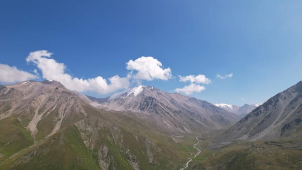 Clouds flow around the tops of high mountains. Rocky gorges and cliffs. The sky is blue, the sun is shining brightly, green grass is growing in places. Mountain terrain from the height of the drone