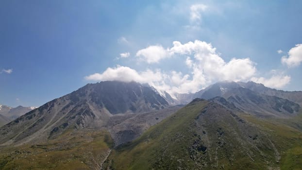 Clouds flow around the tops of high mountains. Rocky gorges and cliffs. The sky is blue, the sun is shining brightly, green grass is growing in places. Mountain terrain from the height of the drone