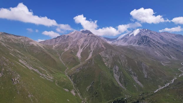 Clouds flow around the tops of high mountains. Rocky gorges and cliffs. The sky is blue, the sun is shining brightly, green grass is growing in places. Mountain terrain from the height of the drone