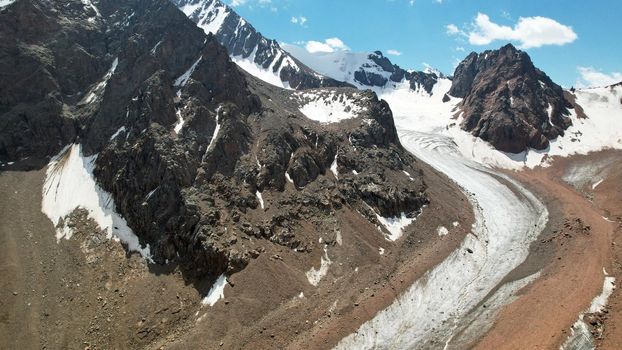 High rocky mountains are sometimes covered with ice and snow. A huge glacier passes between the peaks. The ice is gradually melting. The stones are lying on glacier. Steep cliffs. The view from drone