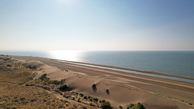 A high sand dune on the ocean shore. Small yellow-green grasses and bushes grow. The blue color of the water. The sun is shining brightly, reflecting in the water. Top view from the drone. Kapchagai