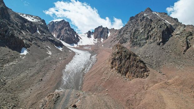 High rocky mountains are sometimes covered with ice and snow. A huge glacier passes between the peaks. The ice is gradually melting. The stones are lying on glacier. Steep cliffs. The view from drone