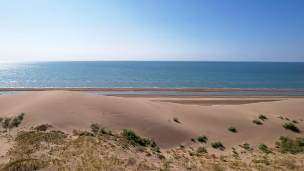 A high sand dune on the ocean shore. Small yellow-green grasses and bushes grow. The blue color of the water. The sun is shining brightly, reflecting in the water. Top view from the drone. Kapchagai