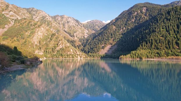 View of the mountain lake Issyk from a height. Turquoise-emerald water from the glacier. Reflection of mountains, forests and the sun on the water. The river flows into the lake. Issyk Dam. Kazakhstan